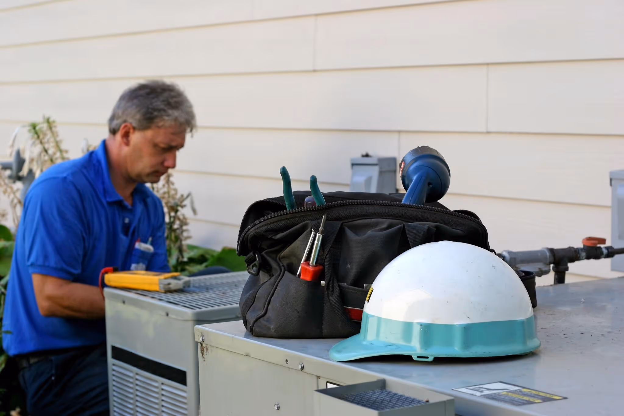HVAC technician working on outdoor unit with tools and white hard hat on nearby surface.