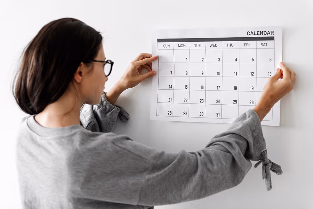 Woman with glasses hanging a printed calendar on a white wall, viewed from the side.
